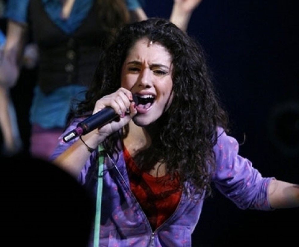 Young Ariana Grande singing with mic and brown curly hair, red top purple hoodie