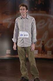 Clay Aiken smiling at his American Idol audition and wearing a striped button down shirt, green pants, and black glasses.