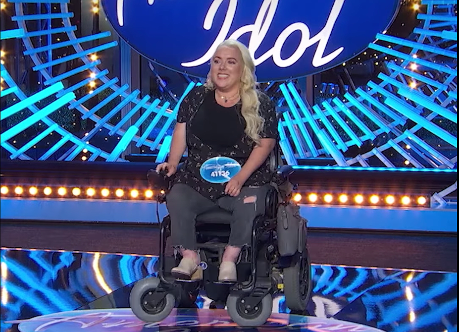 Marna Michele smiling at her American Idol audition, sitting in her black electric wheelchair, and wearing a black floral shirt over a black tank top with ripped jeans.