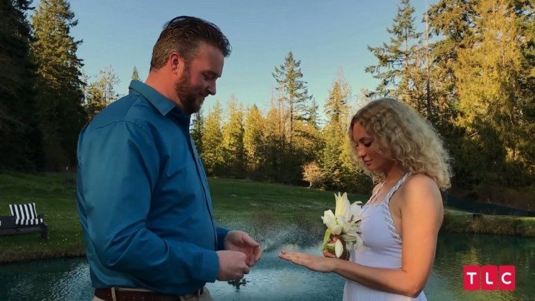 Mike wearing a blue button down shirt and Natalie wearing a white dress while holding flowers at their wedding.