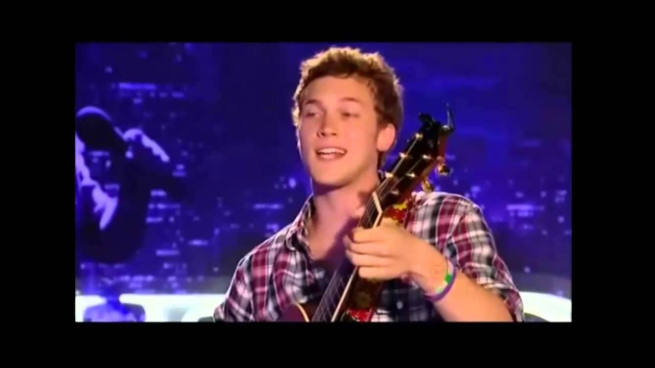 Phillip Phillips singing and playing the guitar at his American Idol audition and wearing a red plaid shirt.