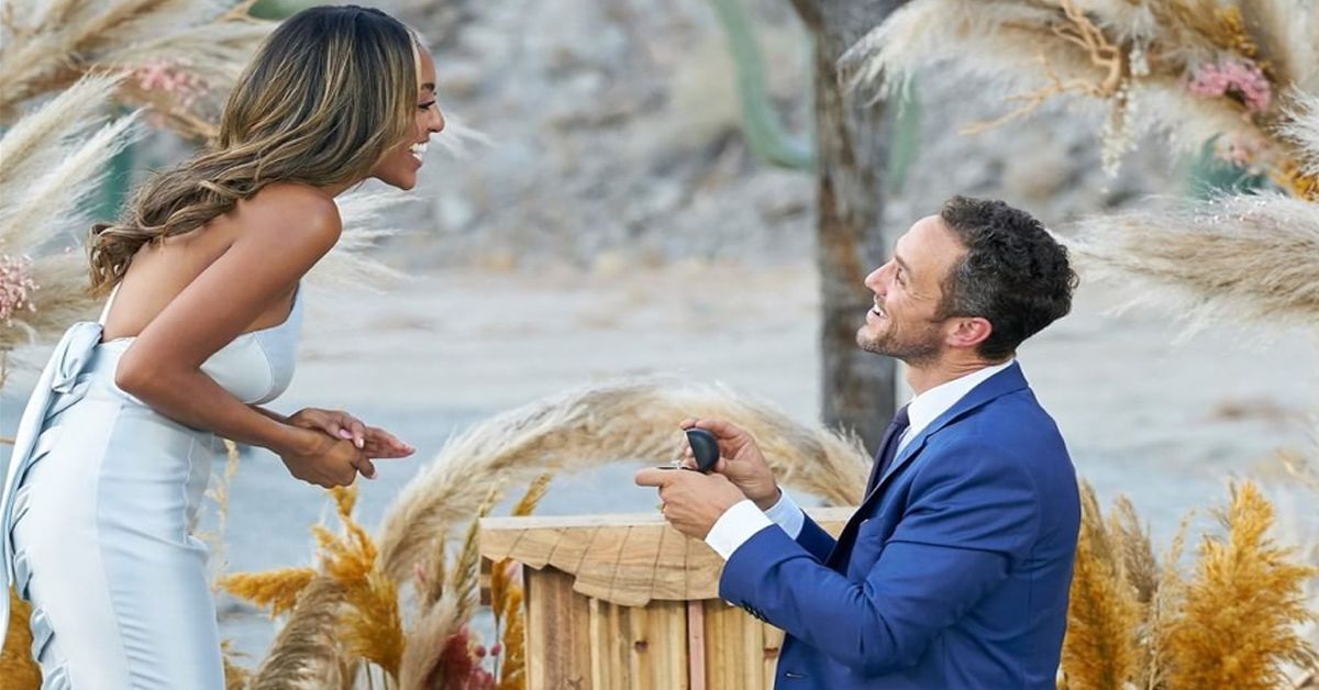 Man in a dark blue suit proposing to the Bachelorette who's smiling and wearing a silver dress.