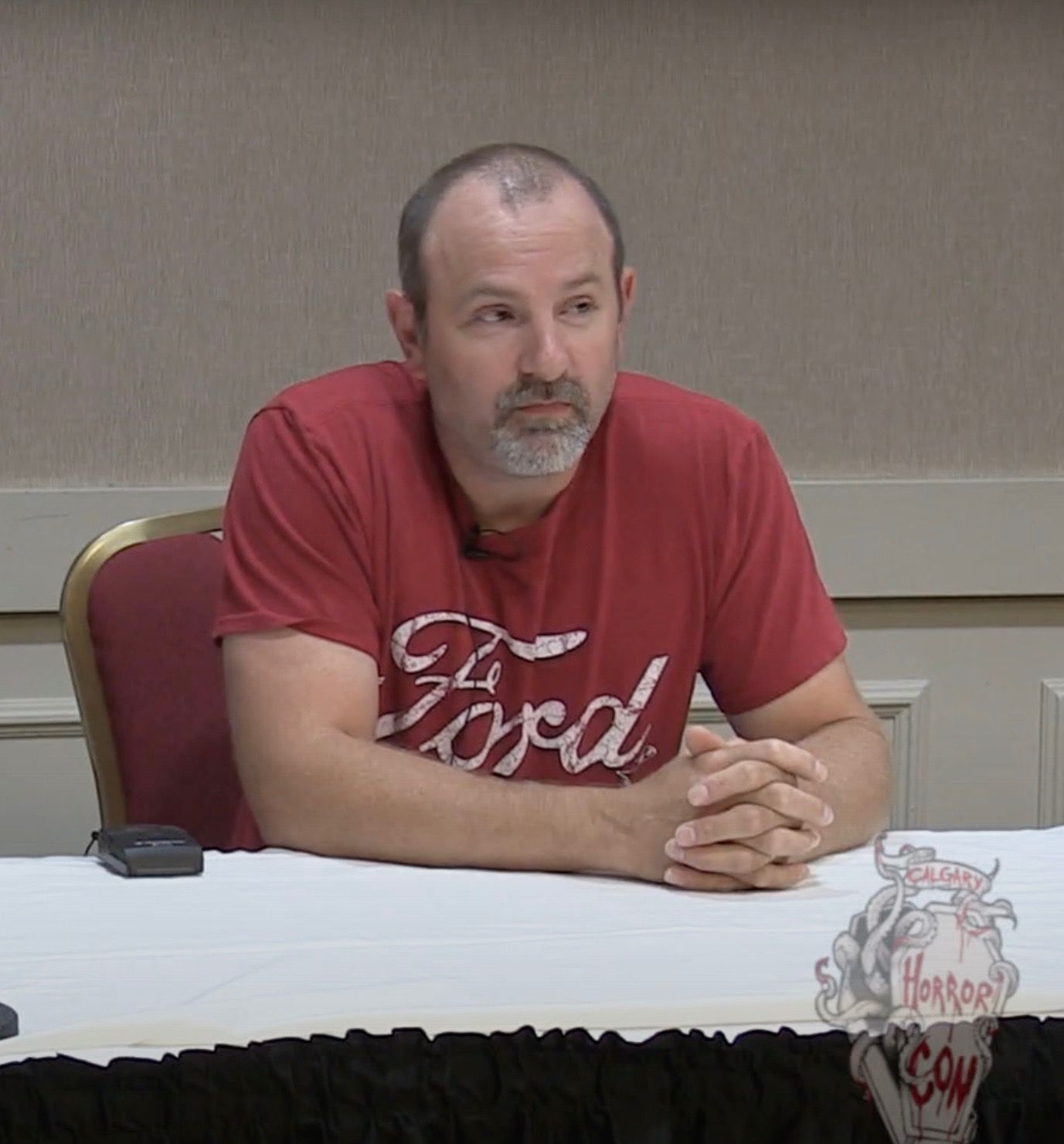 Danny Lloyd wearing a red Ford shirt with his hands on a white table and speaking at a conference.
