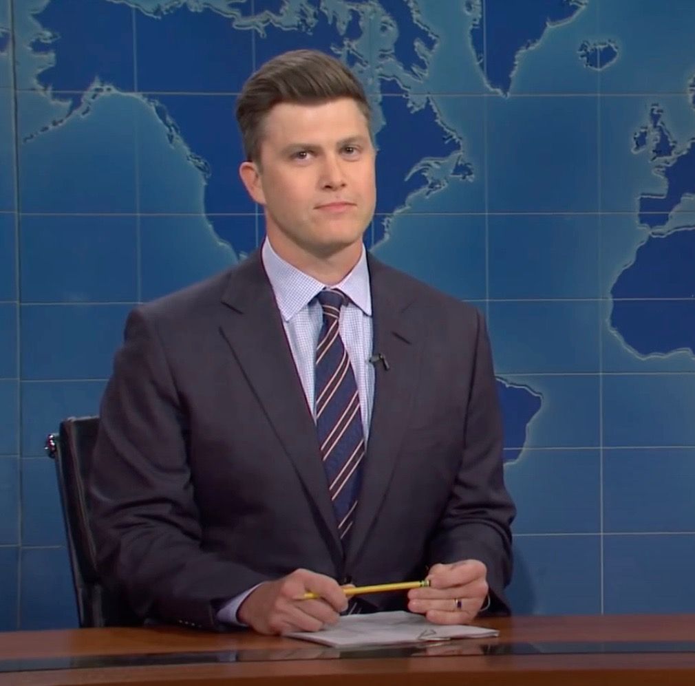 Colin Jost sitting at a news desk on SNL.