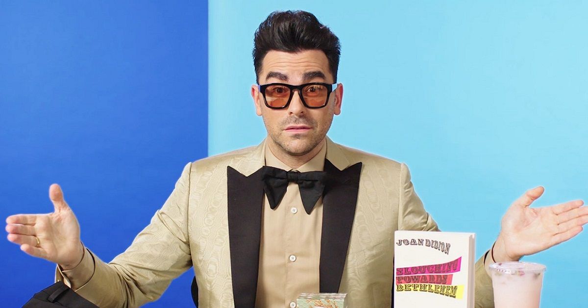 TV star Dan Levy poses in a tan coloured tuxedo and red-tinted sunglasses in front of a blue background