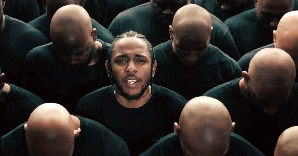 Kendrick Lamar in black shirt among crowd of men in black shirts with their heads down.