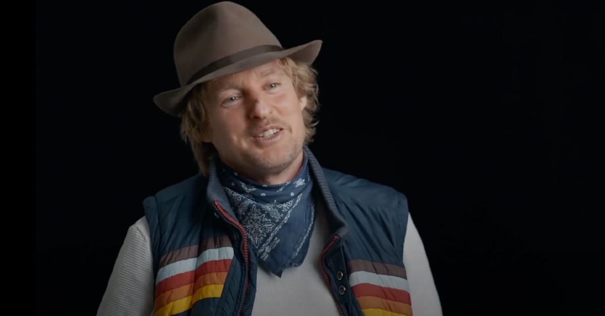 Owen Wilson smiling and wearing a blue bandana around his neck and a tan hat.