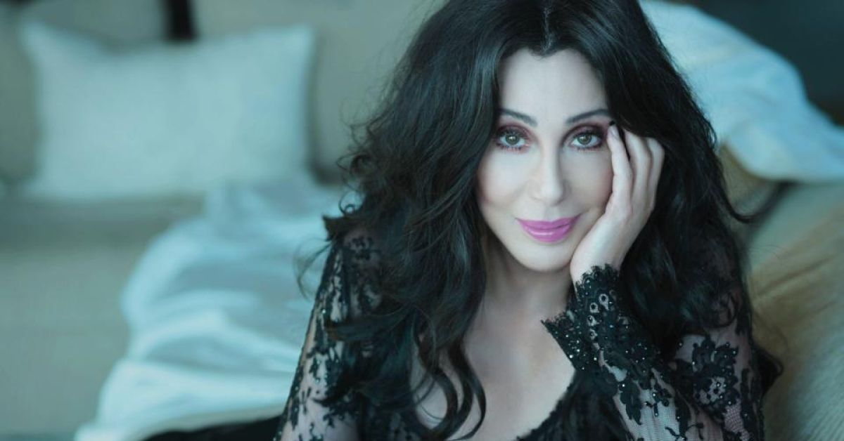 Cher posing in a black lace gown, with a hand holding her cheek and smiling