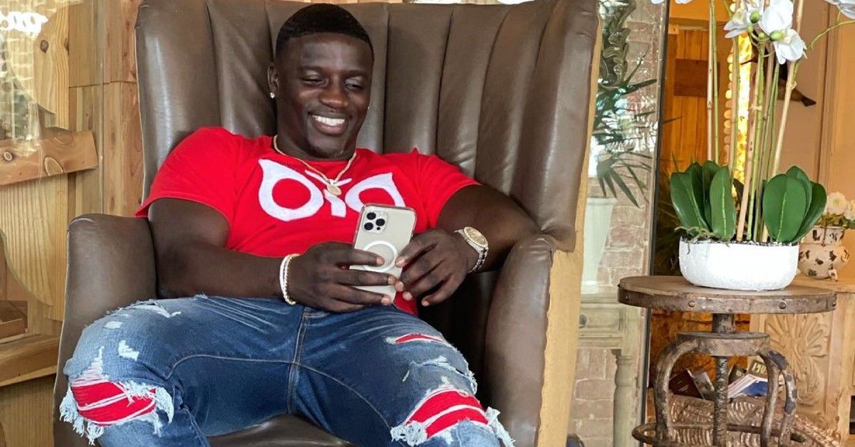 Akon in red shirt and jeans holds phone while sitting in brown chair