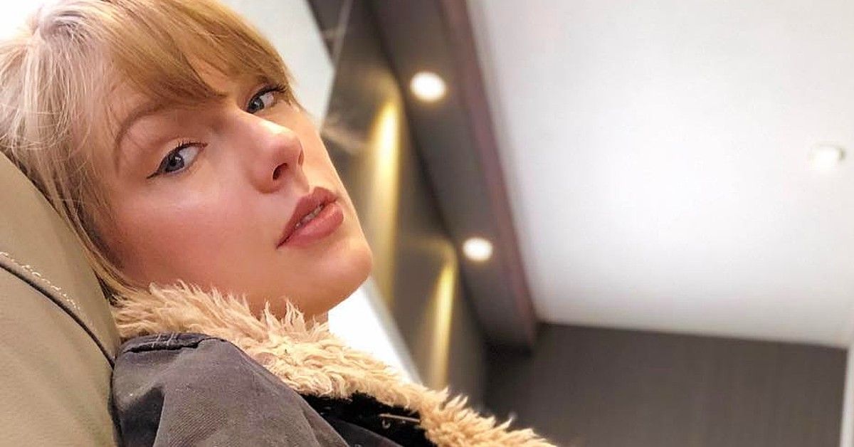 Taylor Swift in black jacket with tan fur sits in trailer alone