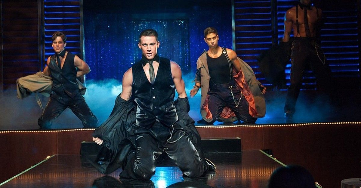 Channing Tatum in black vest and leather pants on stage as Magic Mike with cast behind him 