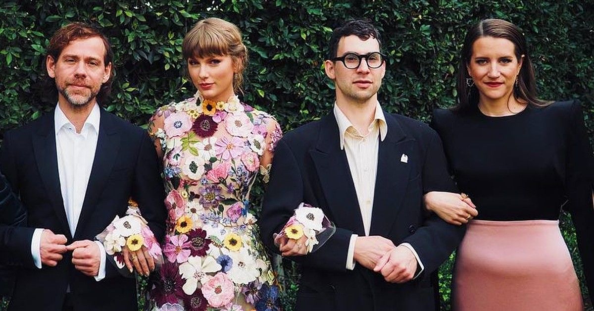 Aaron Dessner in black suit next to Taylor Swift in floral dress with others in front of bush background