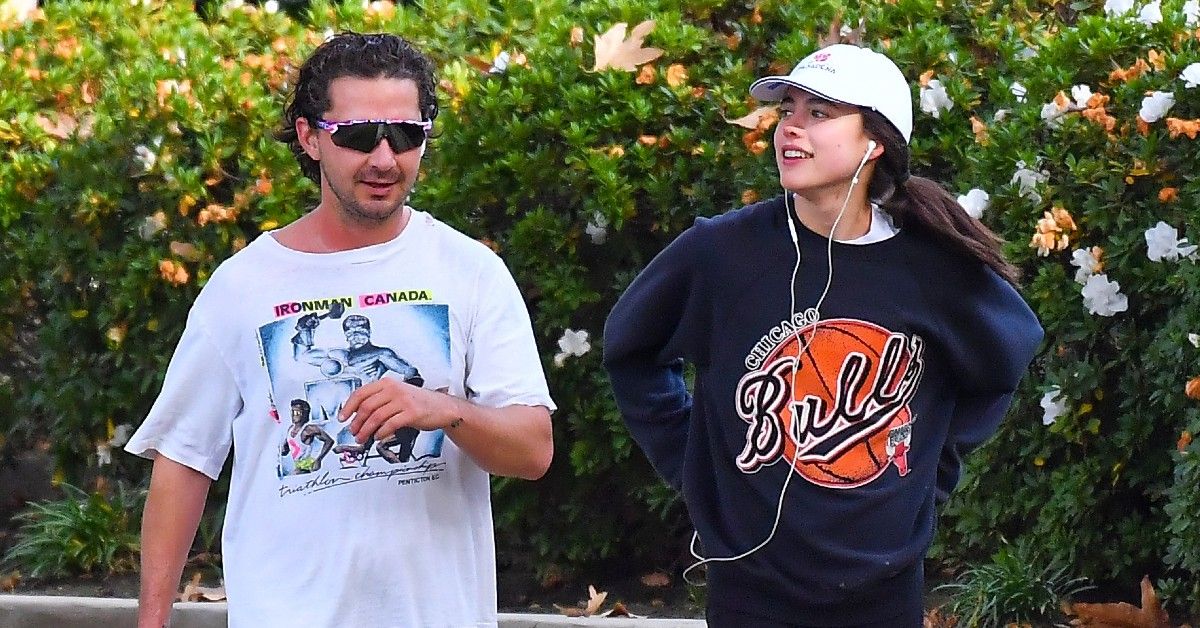 Shia Labeouf in graphic white t-shirt and sunglasses walks outside next to Margaret Qualley in Bulls sweatshirt and white cap