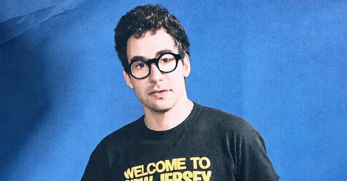 Jack Antonoff in black t-shirt with yellow words stands against blue background 