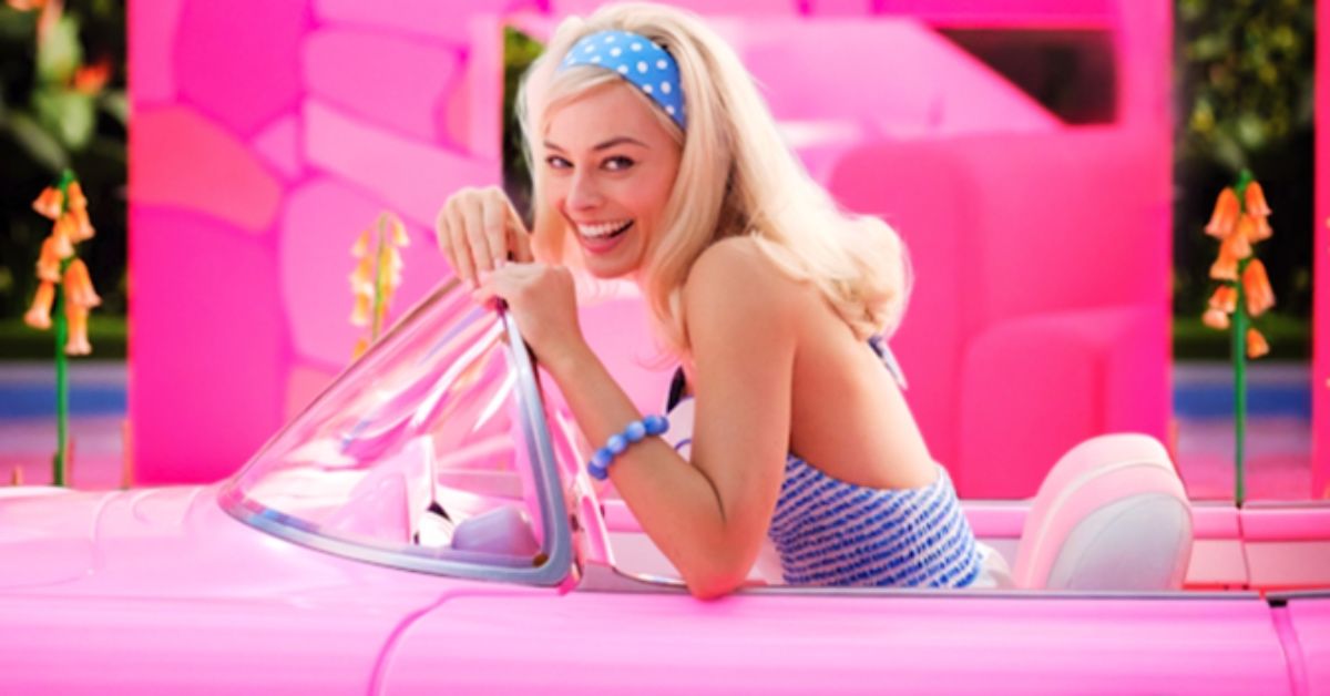 Actress Margot Robbie wears a polka dots headband and a striped blue top as Barbie. She's smiling at the camera, driving a pink convertible car with the roof down.