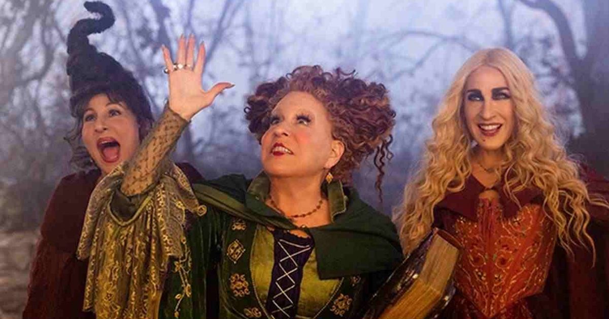 Kathy Najimi, Bette Midler and Sarah Jessica Parker as Mary, Winnie and Sarah Sanderson in Hocus Pocus 2. They seem elated and are looking up at something offscreen as Winnie holds a book under her left arm and raises her right hand.