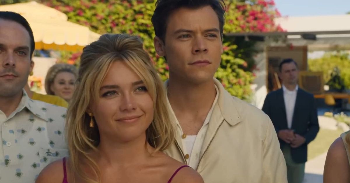 Florence Pugh as Alice and Harry Styles as Jack in a scene of Don't Worry Darling. She is smiling, wearing a spaghetti strap dress and her blonde hair in a half ponytail. He is behind her, looking at someone or something in the distance, wearing a cream jacket and a white, unbuttoned shirt.