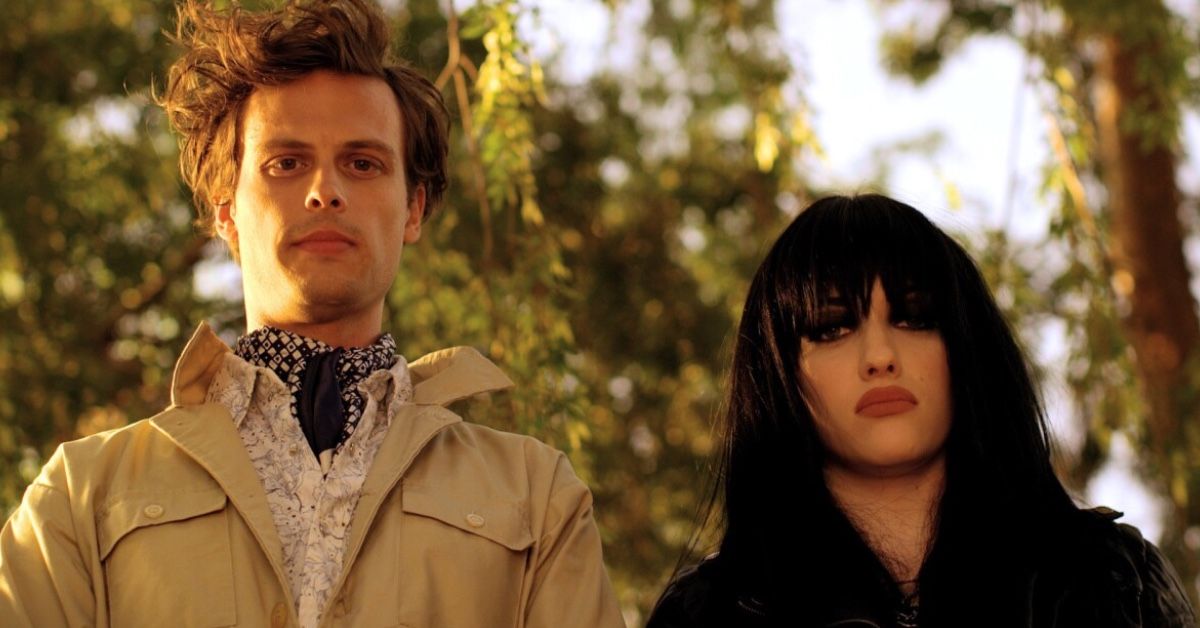 Matthew Gray Gubler and Kat Dennings as Raymond and Becca in a scene of Suburban Gothic. He wears a khaki jacket and a silk scarf while she has dark hair and heavy, gothic makeup.