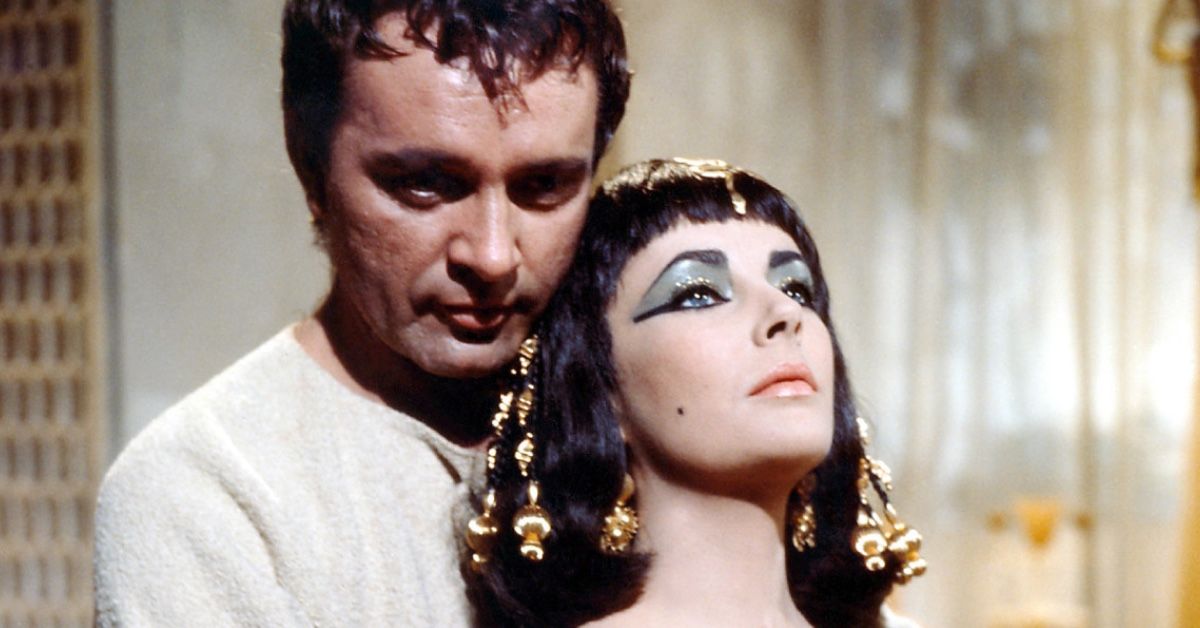 Richard Burton as Marc Anthony and Elizabeth Taylor as Cleopatra in a scene of the 1963 movie of the same name. He's holding her in his arms from behind, as she reclines her head on his chest, looking pensive.