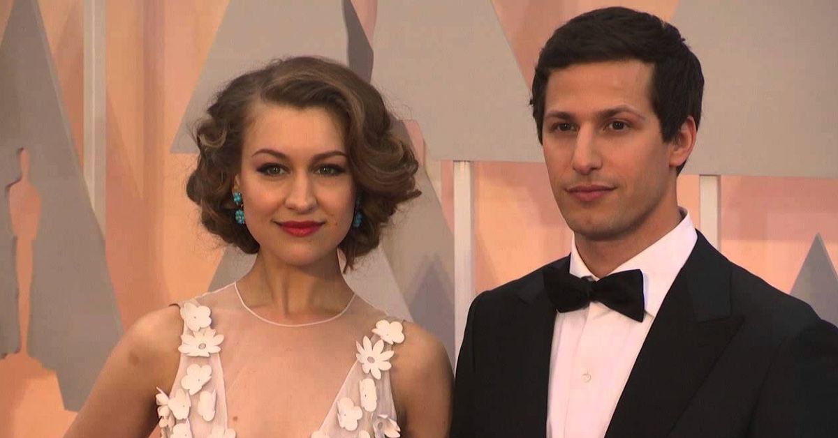 Joanna Newsom and Andy Samberg on the red carpet at the Oscars in 2015. She's wearing a white dress with floral decorations on the sheer bodice and her shoer hair in waves and turquoise earrings, while he's wearing a black suit with a white shirt and a black bowtie.