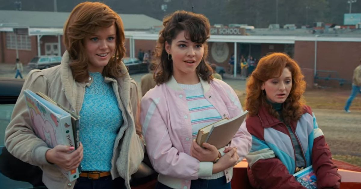 Madelyn Cline (center) as Tina in a scene of Stranger Things. She is with two redheaded friends in the parking lot before class. She wears a light pink jacket and her brown hair in a half ponytail. She's holding some books and notebooks and her attention seems to be caught by something or someone in front of her.