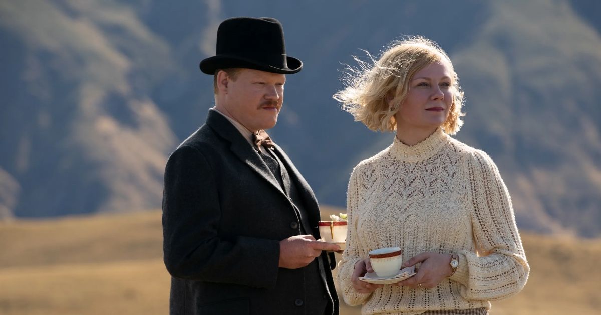 Actors Jesse Plemons and Kirsten Dunst as George Burbank and Rose Gordon in a scene of The Power of the Dog. They're outdoors, it's windy and they are looking in the same direction as they hold one cup of tea each.