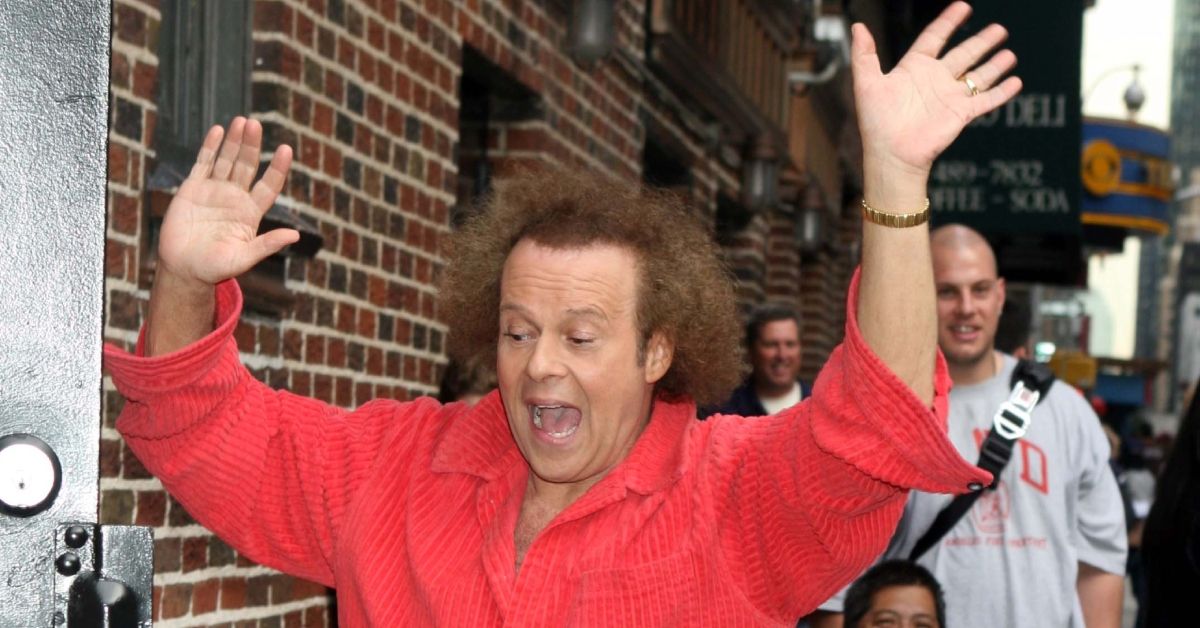 Richard Simmons holding his hands up outside in a red shirt