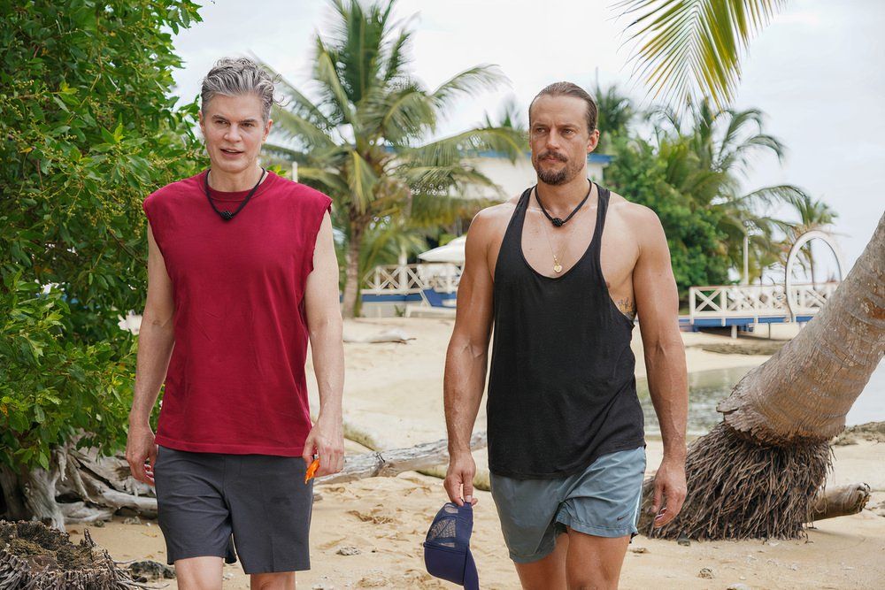 a man in a red sleeveless shirt walks on a beach with a man in a black tank